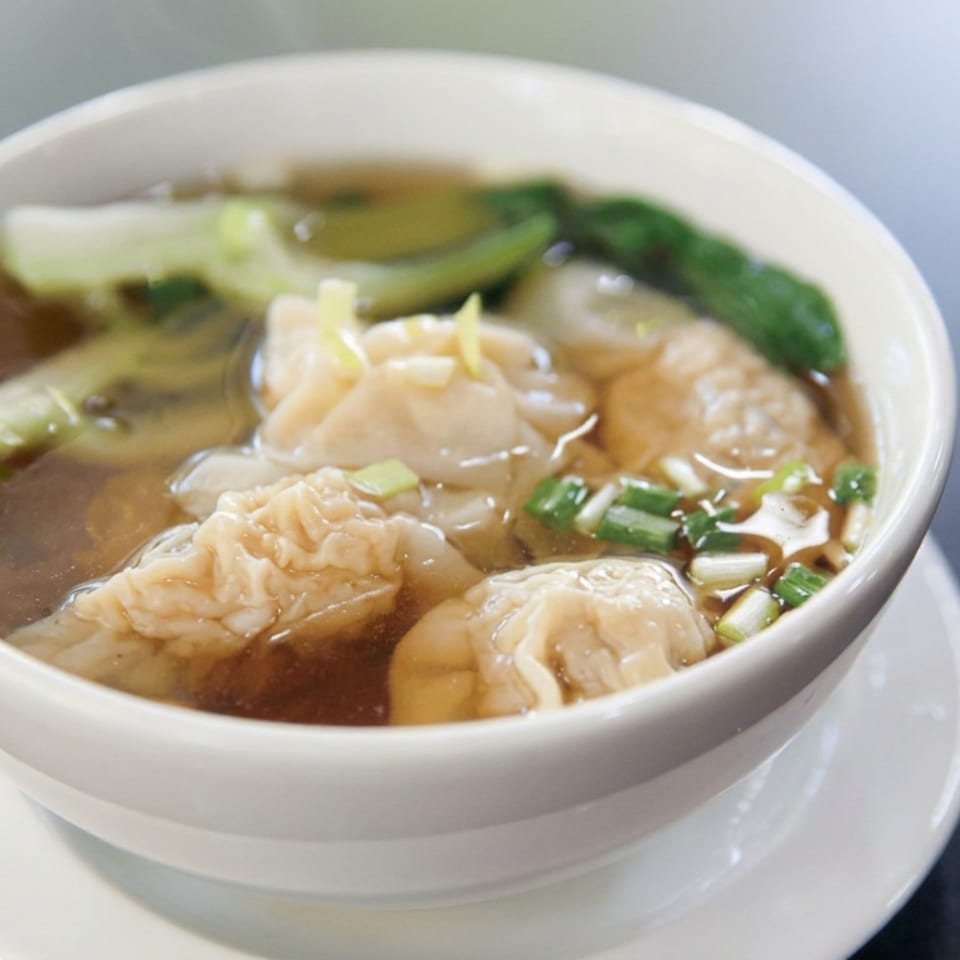 Bowl of clear broth soup with dumplings, leafy greens, tofu, and chopped green onions on a white saucer.